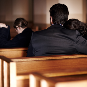 Two pairs of people are sitting in church pews with their backs to the camera. Everyone is wearing black.