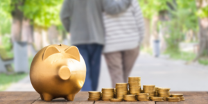 Two people with arms around each other are walking away from the camera and are out of focus. There is a gold piggby bank in the foreground with piles of coins sitting beside it. They may be of the age that allows them to recontribute