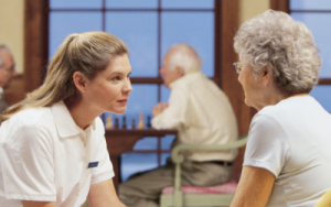 White nurse with a ponytail speaking to older lady. Both women are wearing white shirts. An older man is out of focus in the background.