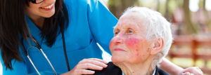 An elderly person with bright pink cheeks is turning towards a care worker or doctor who is wearing blue scrubs. The staff member is smiling and has a stethoscope around her neck