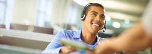 A young man is wearing a blue shirt and a headset with a microphone. He is smiling at someone and handing them a document