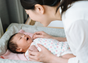 Woman with dark hair leaning over a baby in a polkadot jumpsuit, both are smiling